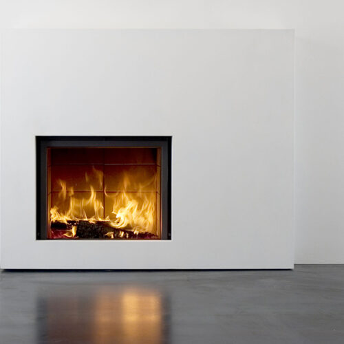 A modern, minimalist stove set into a white wall, with flames visibly burning wood. Its warm glow reflects on the smooth, grey floor.