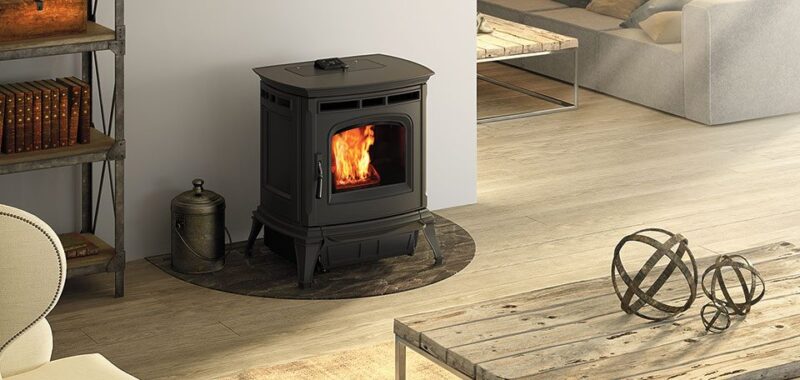 A modern, freestanding black wood stove with visible flames, set in a cozy living room with light wooden flooring, a rustic coffee table, and contemporary furniture.
