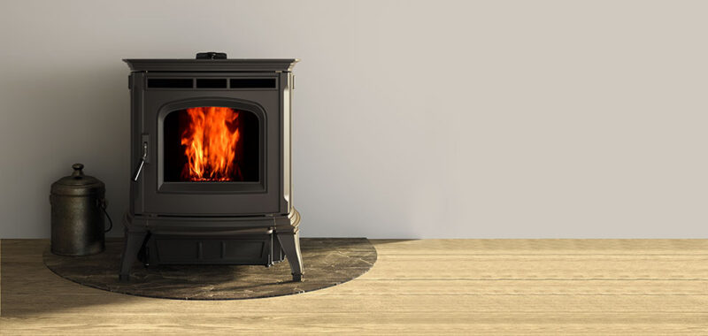 A modern black wood-burning stove with visible flames through the glass door, placed on a wooden floor next to a black metal kettle, against a plain light grey wall.