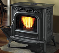 A black cast iron pellet fireplace with intricate designs, featuring a vibrant fire visible through its front glass panel. The stove sits on a beige mat in a room with wooden flooring.