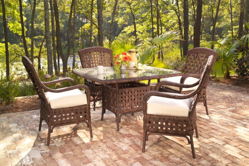 Outdoor patio set with a table, a fire pit, and four wicker chairs on a brick surface, surrounded by lush greenery. The table is set with dishes and a vase of flowers, suggesting
