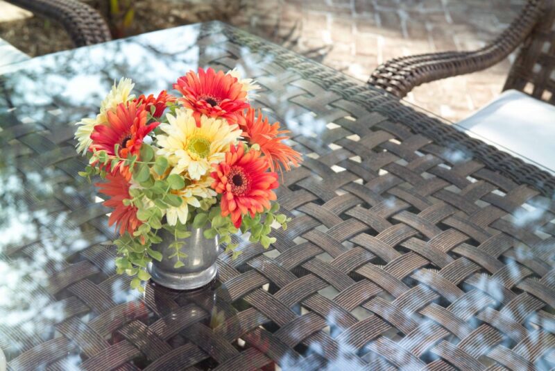 A vibrant bouquet of red and yellow flowers in a metal vase, placed on a glass-top table with a woven dark brown chair near a fire pit in the background, suggesting an outdoor setting.