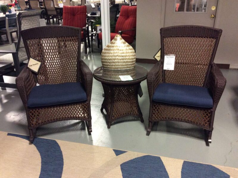 Two brown wicker chairs with blue cushions facing each other, separated by a small wicker table with a textured white vase on it, situated on a patterned carpet near a fireplace in a store setting
