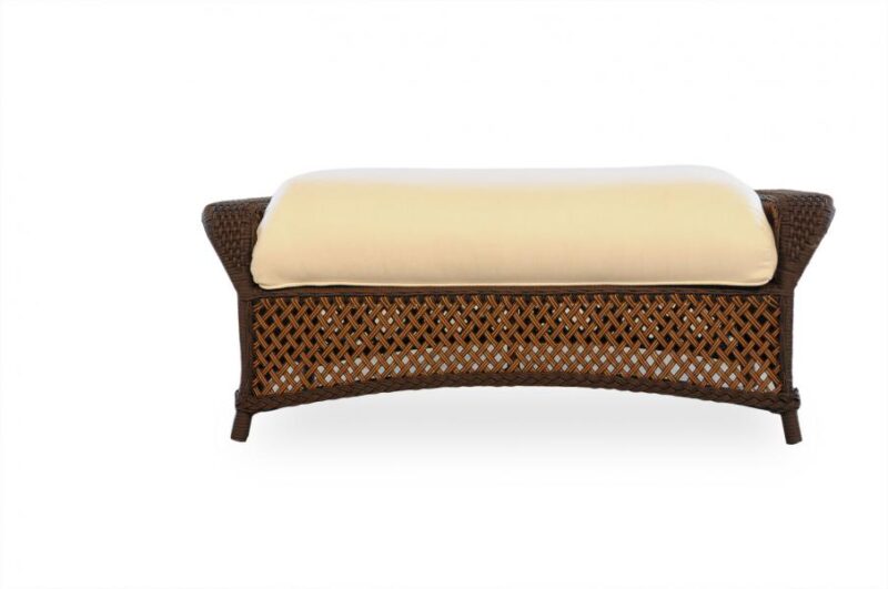 A wicker ottoman with a curved design and dark brown finish, featuring a lattice pattern and topped with a light beige cushion, isolated on a white background. This piece can stylishly compliment an interior
