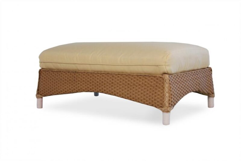 An elegant beige cushioned ottoman with a woven wicker base, featuring short legs and a stove insert, isolated on a white background.