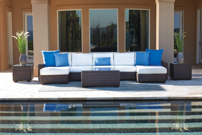 Outdoor poolside setting featuring a modern rattan sectional sofa with white cushions and bright blue pillows, adjacent to a stylish fire pit. The sofa is positioned near a clear swimming pool with a reflection of a