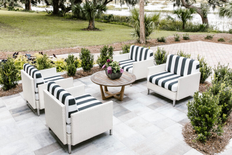 Elegant outdoor seating area with four wicker chairs outfitted in black and white striped cushions, centering around a wooden round table with an integrated fire pit, set against a backdrop of lush greenery