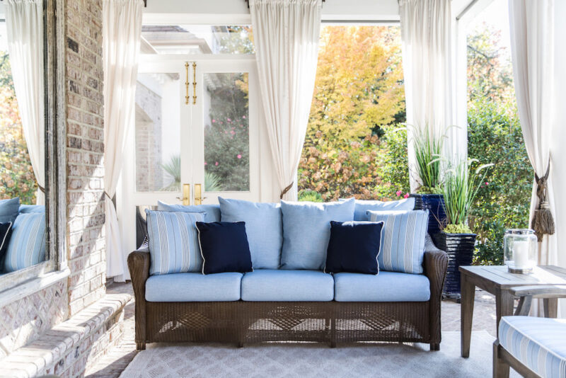 A bright sunroom with transparent curtains and a wicker sofa adorned with blue cushions, overlooking a garden with autumn foliage and a cozy fireplace insert.