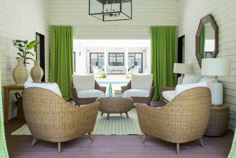 A bright, airy living room with lime green curtains, white furniture, and woven wicker chairs centered around a round wooden table. Large windows and plants add natural light and freshness to the space, enhanced