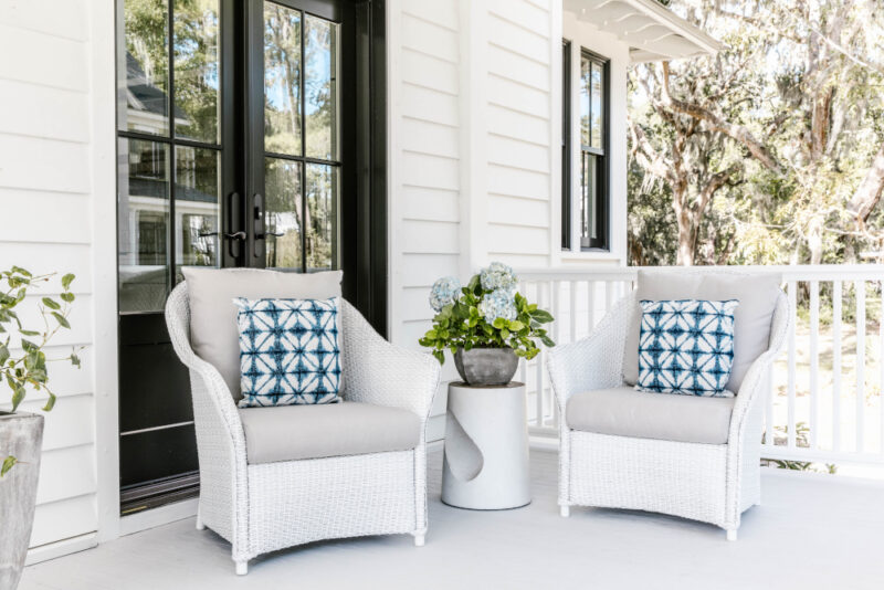Two white wicker chairs with blue patterned cushions on a bright porch, featuring a center table with flowers and a nearby fire pit, near a house entrance surrounded by lush trees.