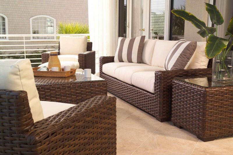 Elegant outdoor patio setup with wicker furniture including a sofa and chairs, cream cushions striped with brown, complemented by a small wooden table, a glass vase with a plant, and an inviting fire