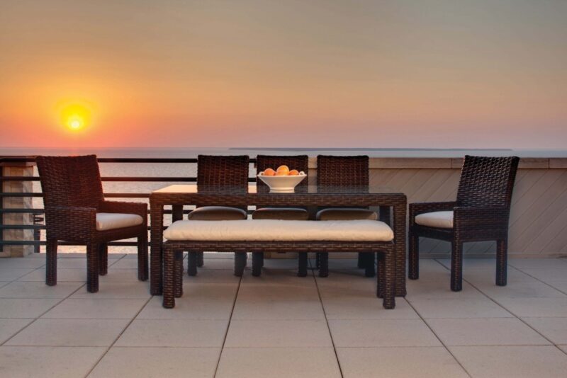 Outdoor dining set on a terrace overlooking the sea at sunset, featuring a wooden table, four chairs with cushions, a bowl of fruit on the table, and a fire pit nearby.