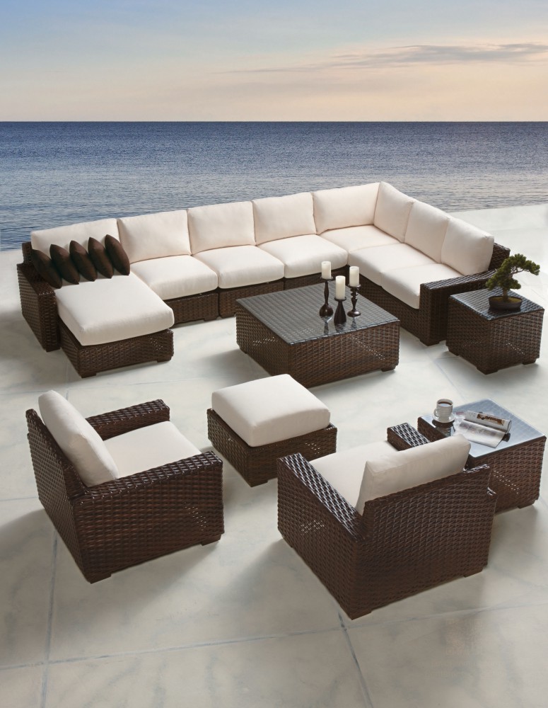 Luxurious outdoor brown wicker sectional sofa with white cushions and matching ottomans around a fire pit on a patio overlooking a scenic beach at dusk.