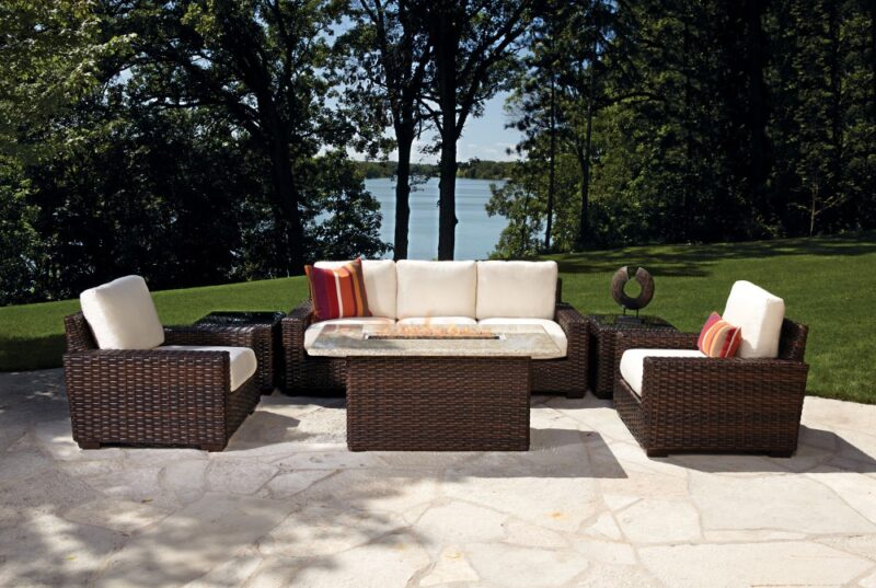 Outdoor patio furniture set including a sofa and chairs with cushions, arranged on a stone pavement, overlooking a tranquil lake surrounded by trees, featuring an insert fire pit.