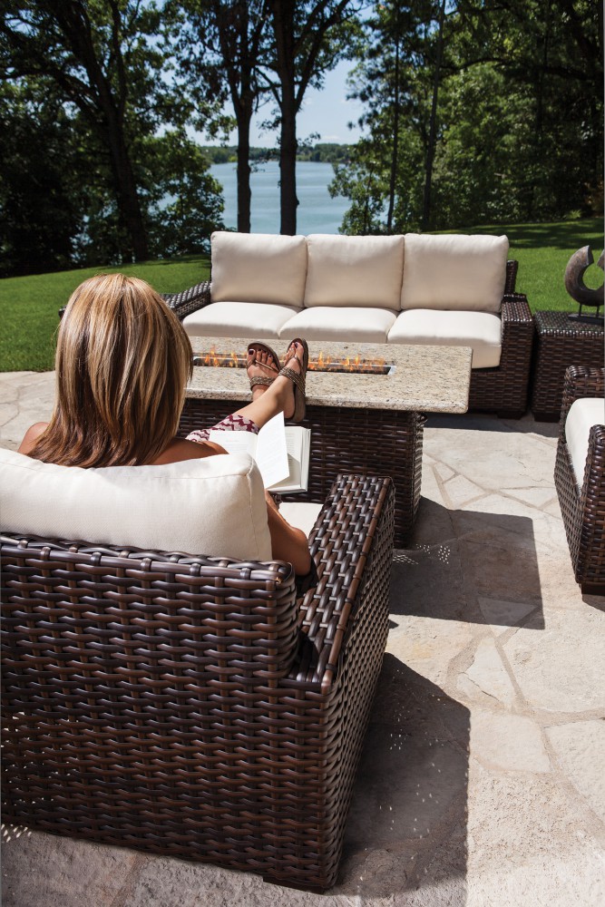 A woman relaxing on a wicker sofa with her feet up overlooking a lake on a sunny day, surrounded by similar patio furniture, a fire pit, and lush greenery.