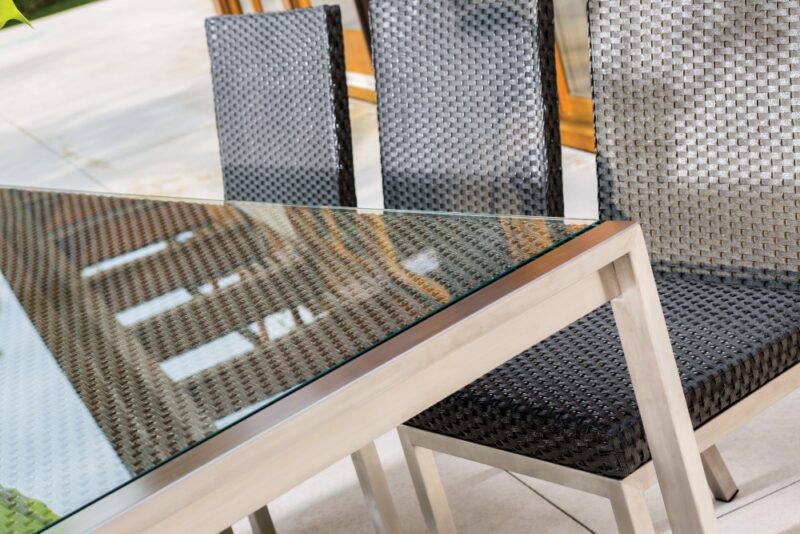 Close-up of a modern outdoor dining set with a glass table and four woven black chairs on a patio near a fireplace.