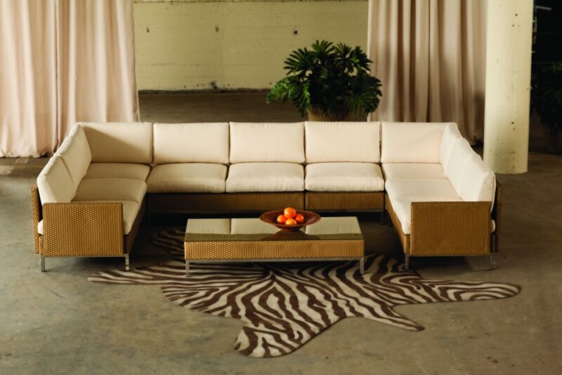 A modern sectional sofa with white upholstery and rattan side panels, in front of beige curtains, near a fireplace, and a coffee table bearing a bowl of oranges, set on a zebra print rug