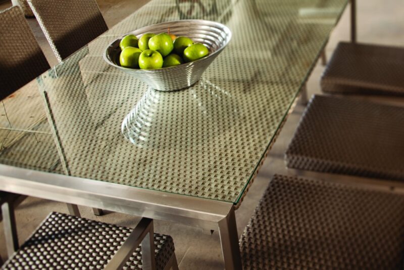 A modern glass table with a textured metallic base, holding a bowl of green apples. Matching chairs with textured seats surround the table, and a fire pit insert nearby enhances the ambiance.