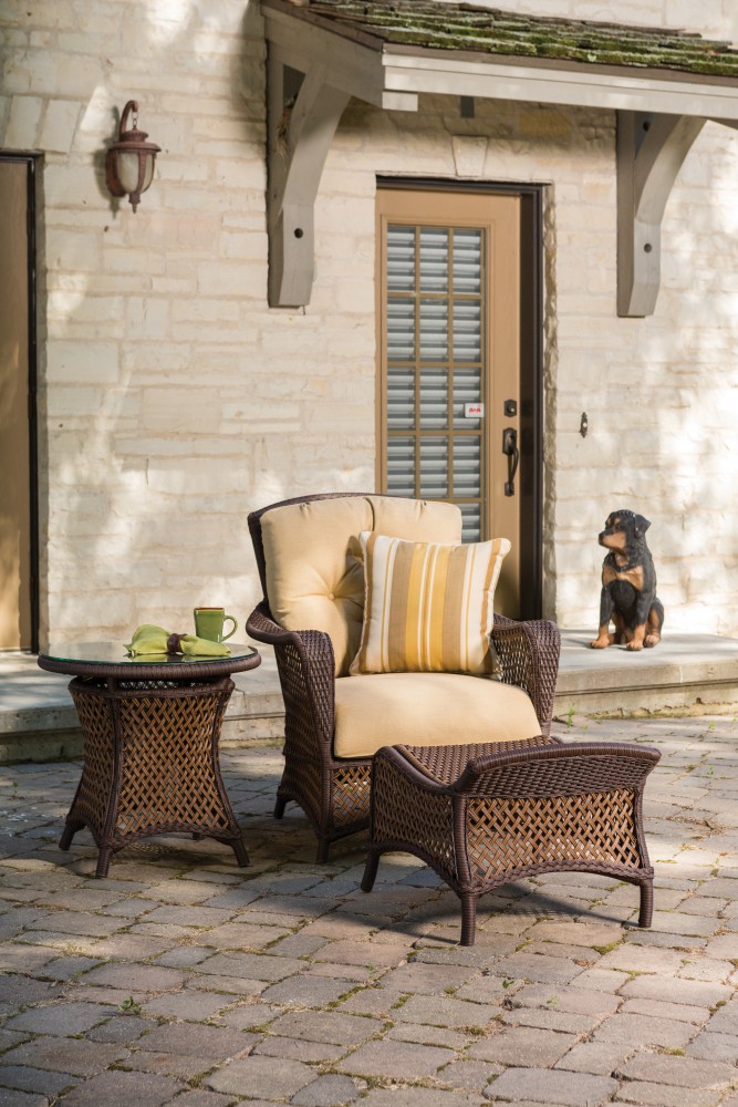 An inviting outdoor seating area with a plush armchair and a small table, next to a stone house and under daylight. A decorative dog statue sits by the door, near a cozy fire pit.