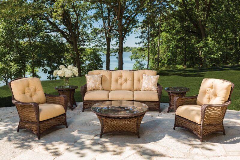 Luxurious outdoor patio furniture set on a stone platform overlooking a calm lake, surrounded by lush trees and featuring a fire pit, on a sunny day.
