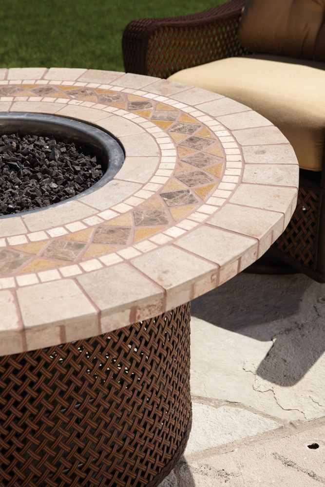 Round outdoor fireplace with a detailed brick and mosaic design, surrounded by a wicker base, next to a plush, beige outdoor sofa on a stone patio.