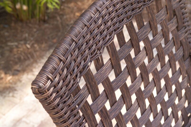 Close-up image of a brown wicker chair focusing on its intricate woven pattern and texture, with a softly blurred background suggesting an outdoor setting near a fire pit.