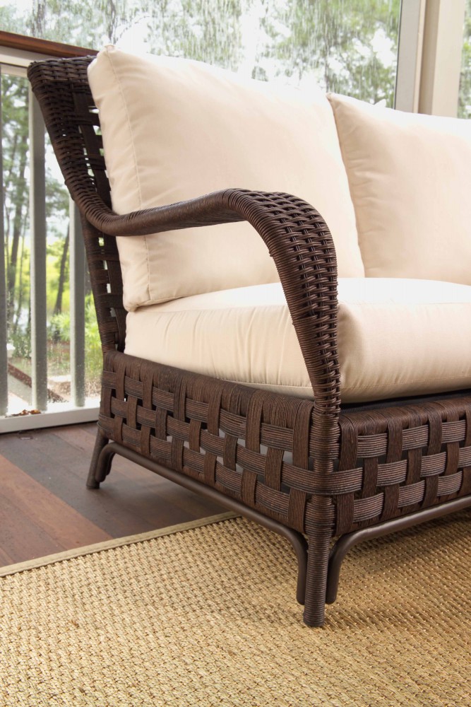 Close-up of a brown wicker armchair with cream cushions on a beige woven rug, next to a large window with a view of greenery outside and an elegant fireplace nearby.