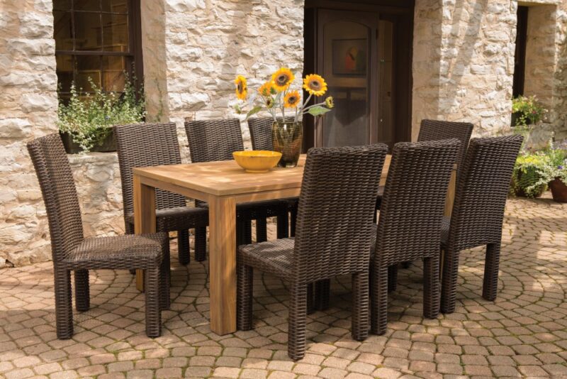 A rustic outdoor dining setup with a wooden table and eight woven chairs on a cobblestone patio. A bowl with sunflowers adds a bright touch next to a stone house with a fire pit.