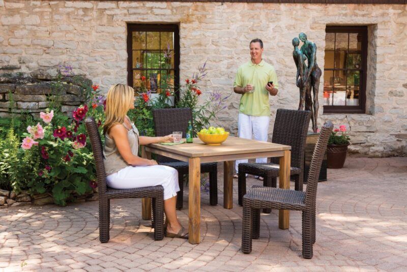 A man and a woman are enjoying a sunny day on a patio. The man is standing with a drink while the woman sits at a table with fruits. A parrot statue, blooming flowers,