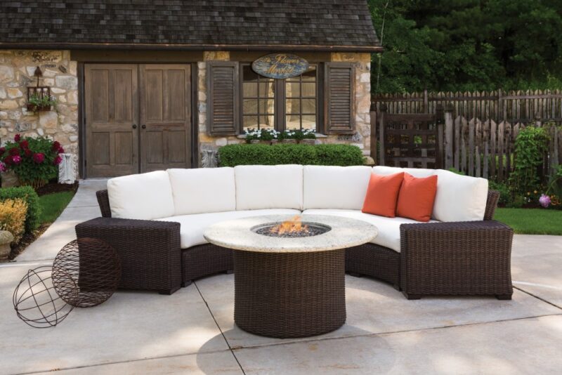 Outdoor living space featuring a curved wicker sectional sofa with white cushions around a round fire pit table, set against a rustic stone house with lush plants and an inserted stove.