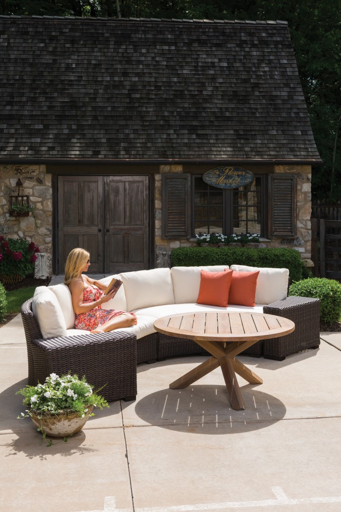 A woman relaxes on an outdoor l-shaped sofa, reading a book in front of a quaint stone cottage with flowers and a lush garden under clear skies. Nearby, an inviting fire pit adds warmth to