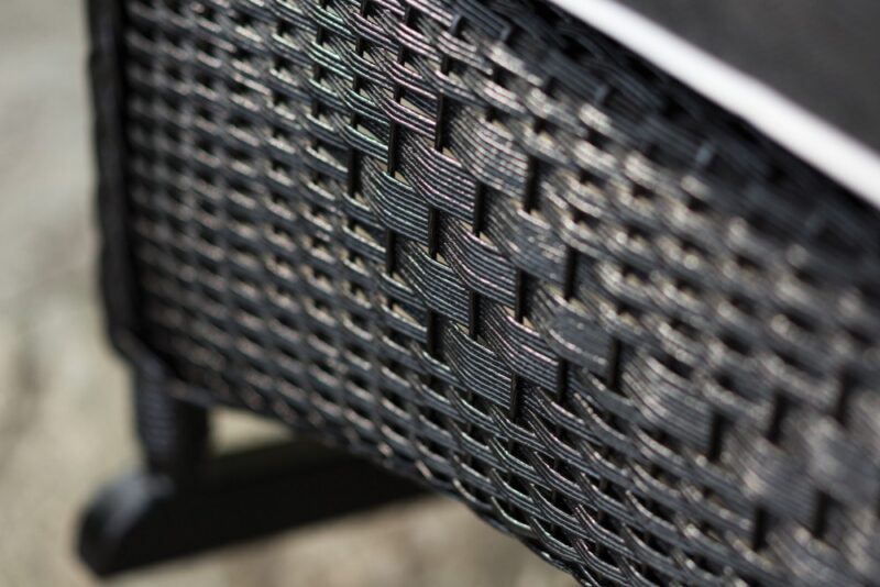 Close-up photo of a detailed, textured black wicker pattern on outdoor furniture, focusing sharply on the weaves with a blurred background featuring a fireplace insert.