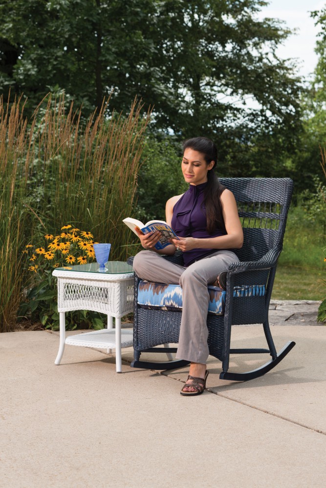 A woman sits in a rocking chair outdoors, reading a book next to a small white table with a coffee cup, surrounded by lush greenery and flowers, near a cozy fire pit.