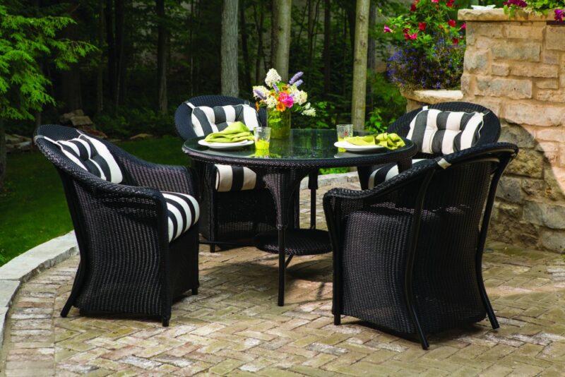 An outdoor dining setup with a round black table and four woven chairs, striped cushions, set on a brick patio surrounded by lush greenery, flowers, and a fire pit.