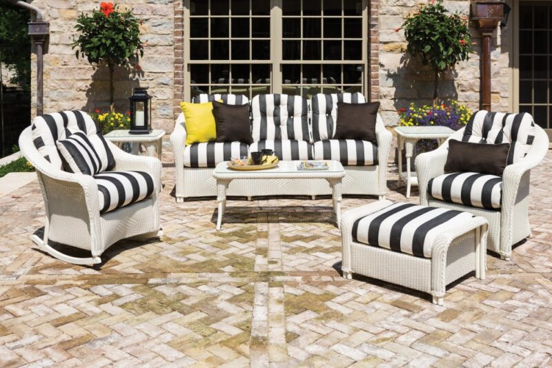 Outdoor patio setting with striped black and white cushions on white wicker furniture, including a sofa, chairs, and a coffee table with a stove insert, on a cobblestone surface. Flowers and a