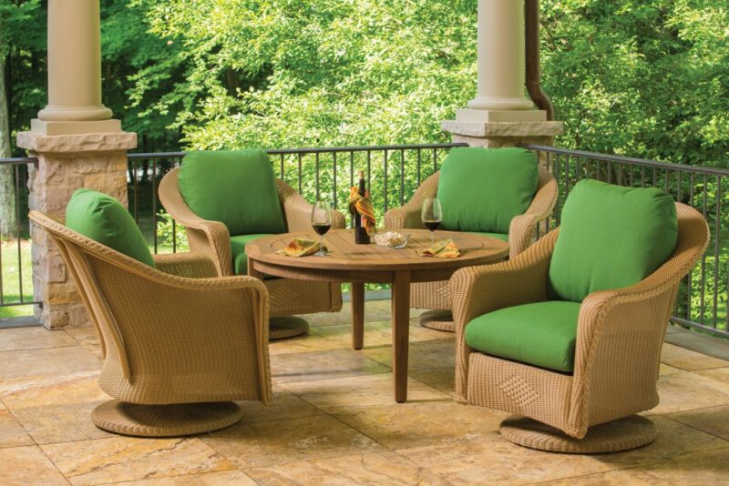 An inviting outdoor patio with wicker furniture, green cushions, and a table set with snacks and wine, surrounded by greenery and forest views, featuring a cozy fire pit.
