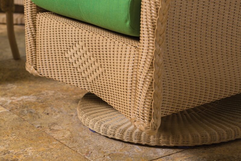 Close-up of a wicker chair with a green cushion, showcasing the intricate weaving and texture, set against a stone tiled floor by a fireplace.