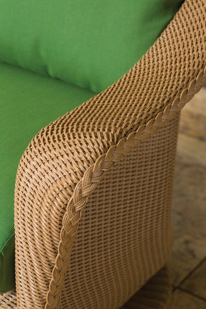 Close-up of the textured arm of a wicker chair with a braided design, complemented by a green cushion and positioned near a fireplace.