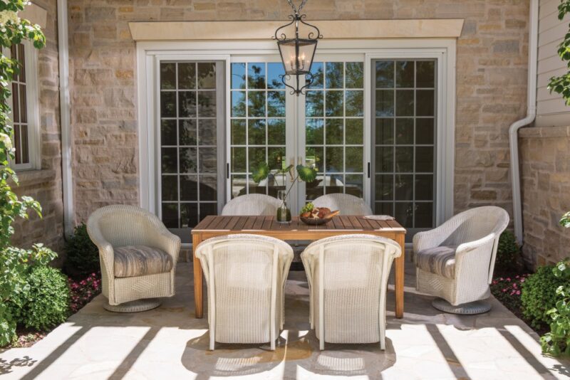 A cozy outdoor patio seating area with a wooden dining table, four chairs, and vibrant plants, against a backdrop of French doors and a stone wall featuring an integrated fire pit.