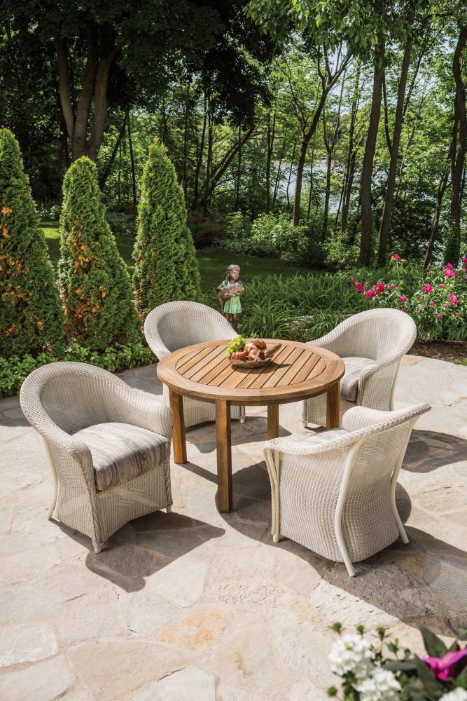 A picturesque outdoor setting with four wicker chairs around a wooden round table, set on a stone patio surrounded by lush greenery and blooming flowers, under a bright, sunny sky. A cozy fireplace