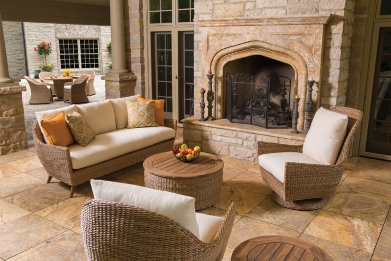 An elegant patio with rattan furniture including a sofa and chairs, arranged around a coffee table. A stone fire pit and dining setup in the background enhance the cozy, outdoor living space.