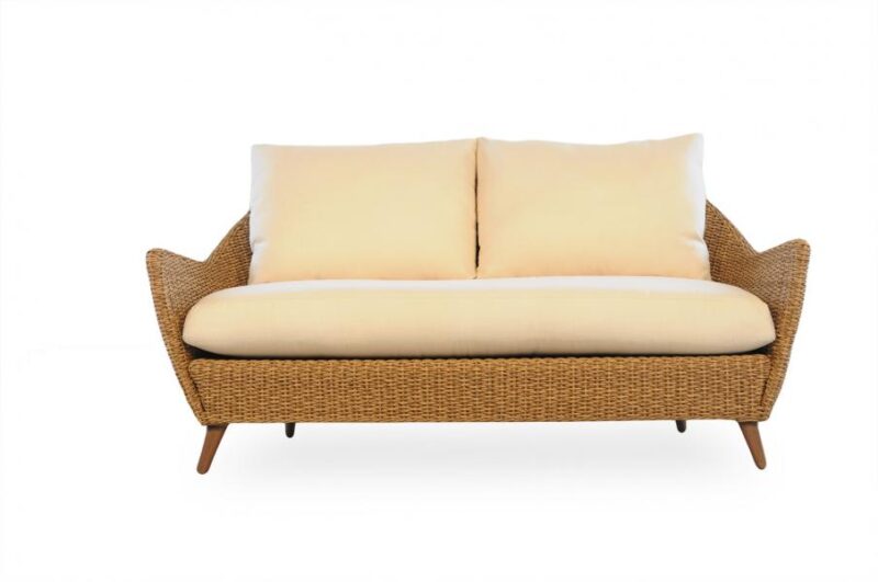 Modern wicker loveseat with a beige cushion and two side pillows near a fireplace, isolated on a white background.