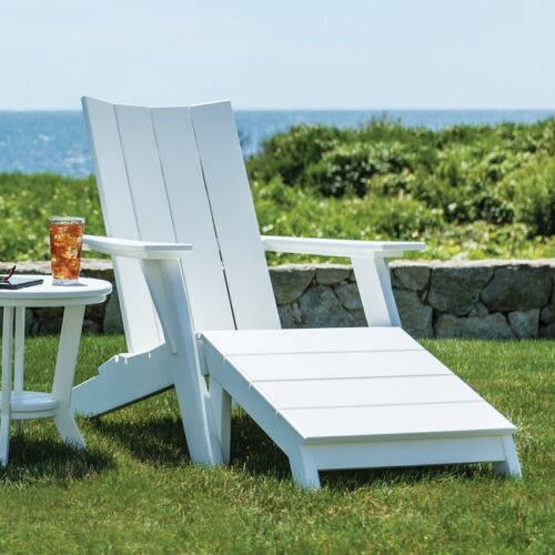 An adirondack chair and a side table with a glass of iced tea, situated on a lush lawn overlooking a tranquil, sunny coastal landscape with a fire pit.