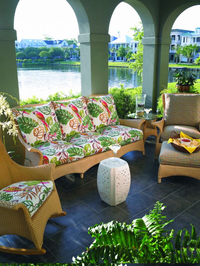 An inviting patio with wicker furniture, including a patterned sofa and chair, set under archways overlooking a serene river. A cozy fire pit enhances the tranquil, relaxing outdoor setting with lush plants.