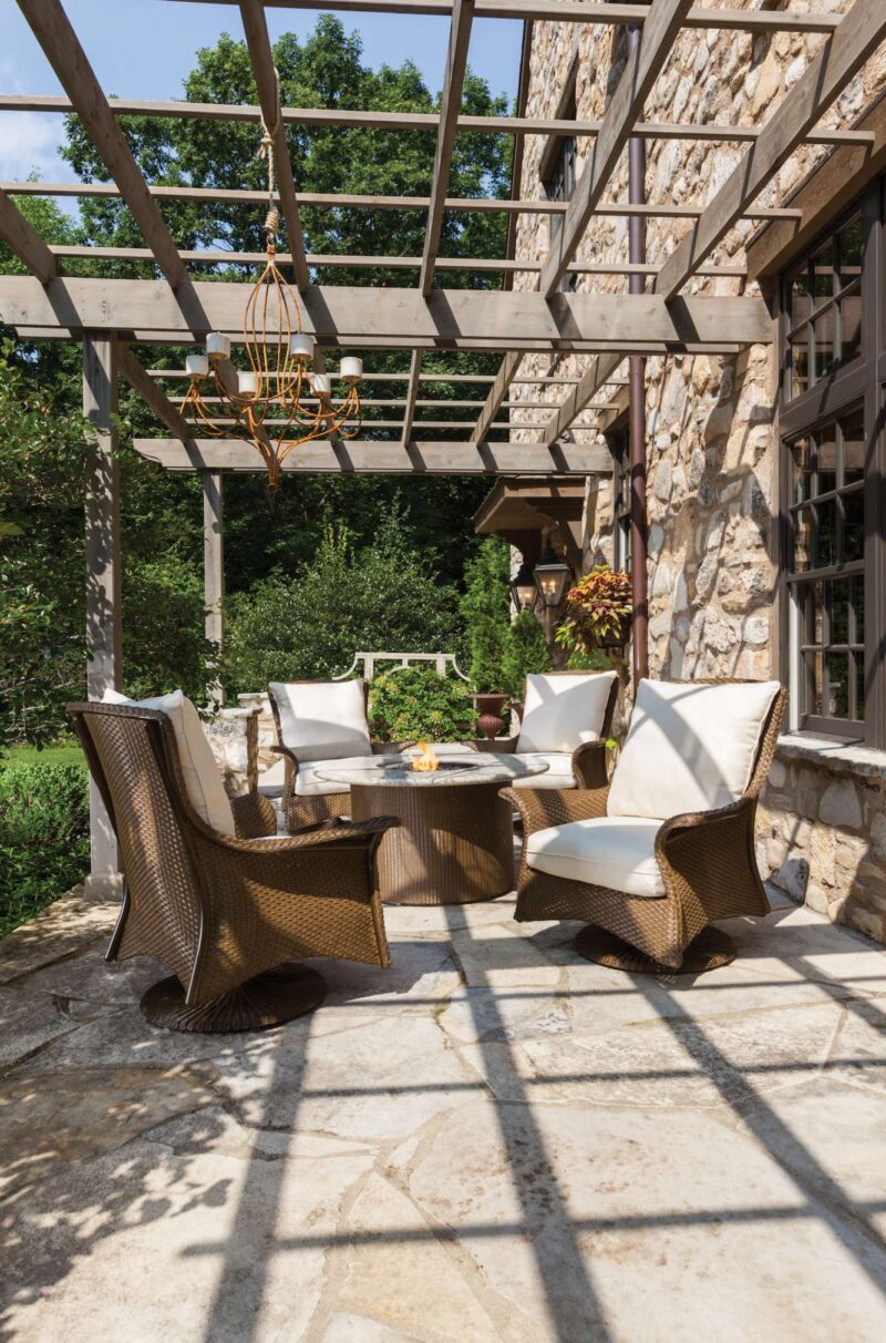 An elegant outdoor patio featuring wicker furniture with white cushions, set on a stone floor under a wooden pergola, with lush greenery and an insert of a stone fireplace in the background.