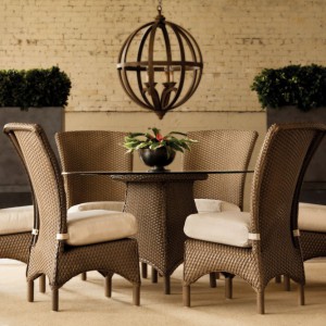 Elegant outdoor dining area featuring a round glass table with four woven rattan chairs and a fire pit, under a spherical chandelier, against a brick wall backdrop.