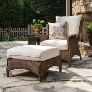 Two brown wicker chairs with white cushions and a matching ottoman on a patio, near a potted plant and a stone wall, with a wooden fence in the background and a fire pit.