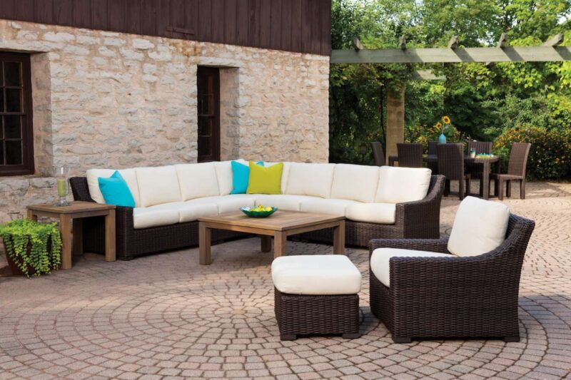 A stylish outdoor patio with a large sectional wicker sofa adorned with white and turquoise cushions, a matching armchair, and a coffee table, set against a rustic stone wall and cobblestone floor,