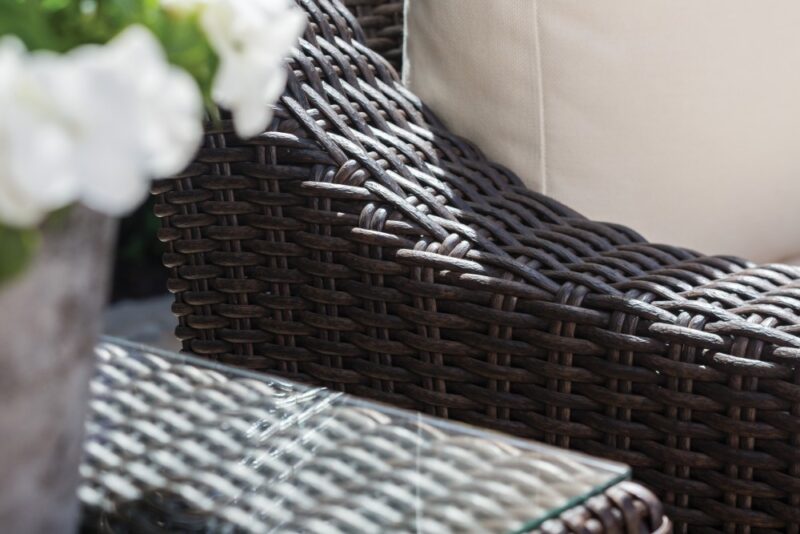 Close-up view of a woven dark brown rattan chair arm next to a glass-top table with a stove nearby, featuring a white cushion and blurred white flowers in the foreground.