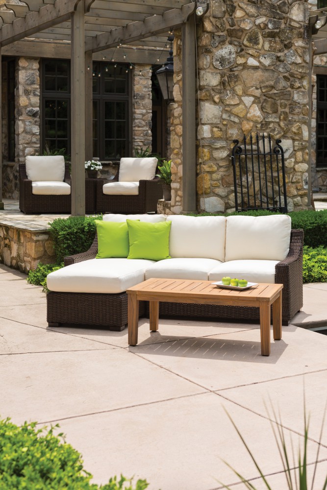 Outdoor patio with stone flooring and a wicker sofa adorned with white and lime green cushions, flanked by two wicker armchairs. A wooden coffee table is in the center, accompanied by a